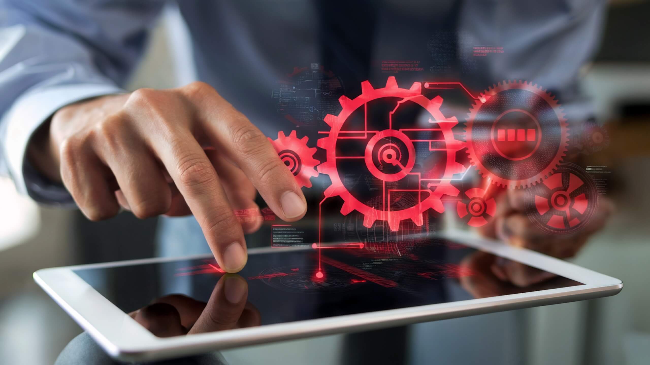 Hand of a person using a tablet, with red gears and technical icons projected on the screen, symbolizing digital processes and automation.