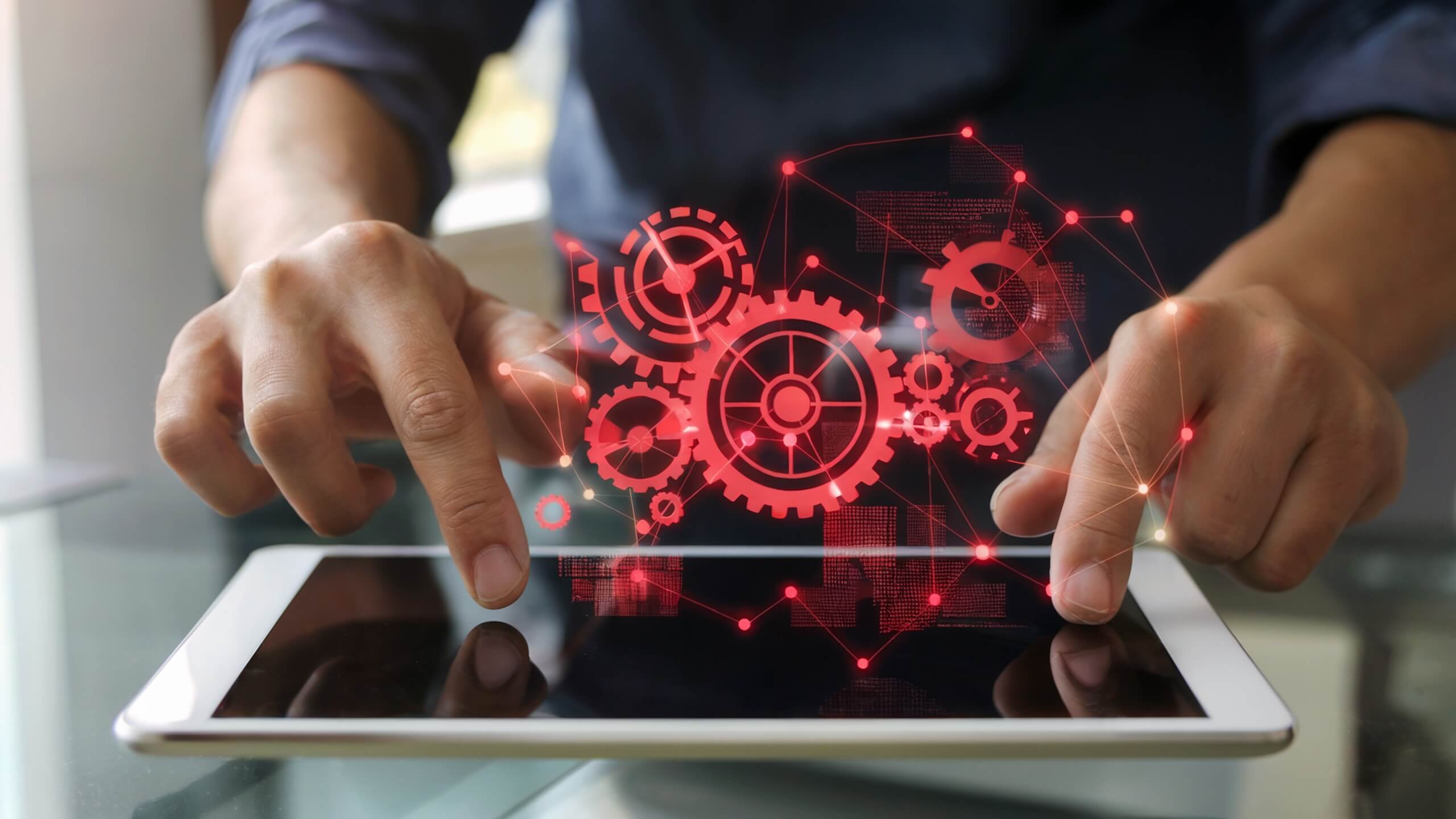 Industrie: Machen Sie Ihren Job - wir machen unseren Zwei Hände, die ein Tablet bedienen, mit roten Zahnrädern und technischen Symbolen, die auf den Bildschirm projiziert werden und digitale Prozesse sowie Automatisierung darstellen.