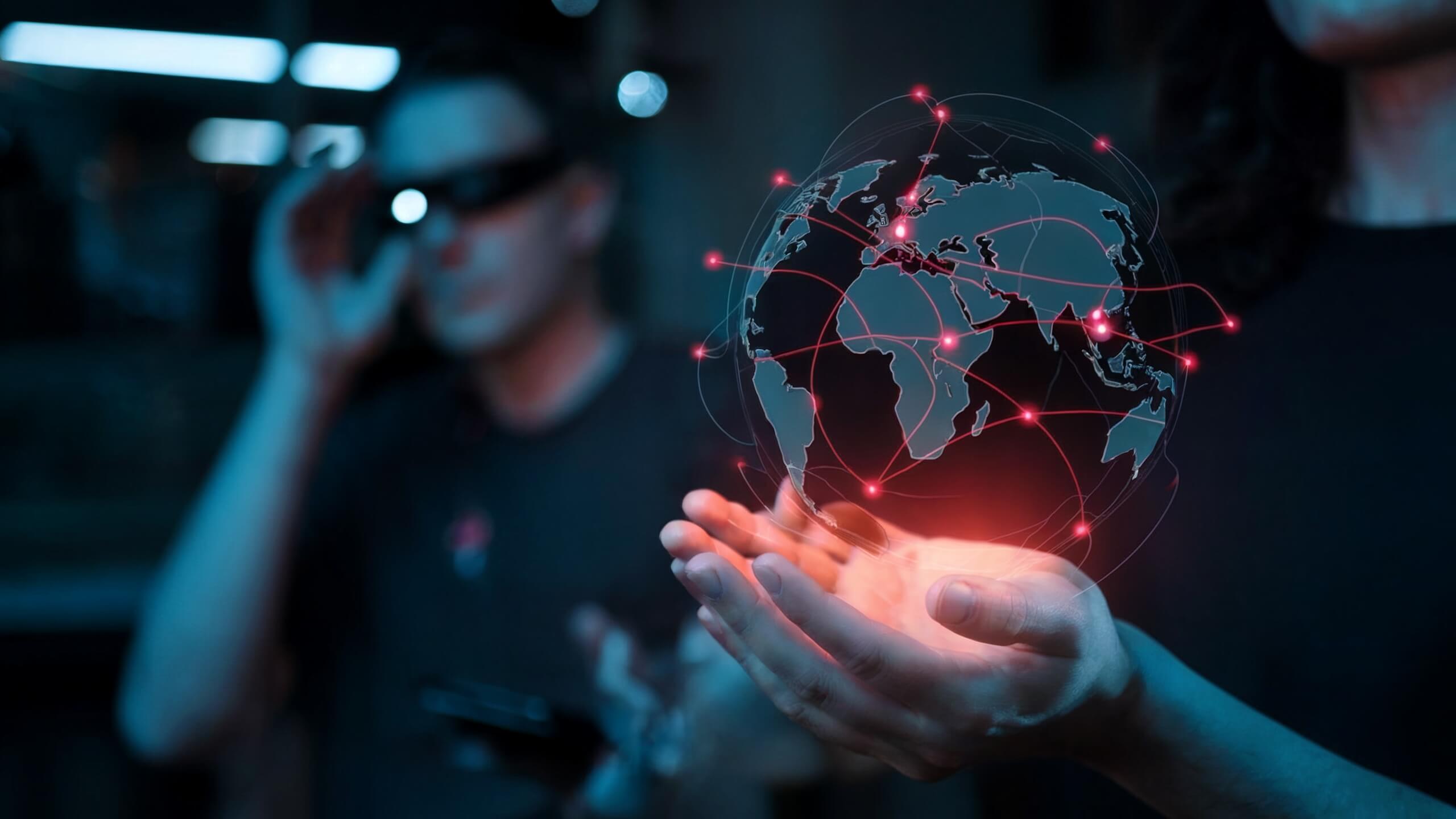 Hände halten eine digitale Weltkugel mit roten Verbindungslinien, die globale Vernetzung symbolisieren, während im Hintergrund eine Person mit AR- oder VR-Brille steht.